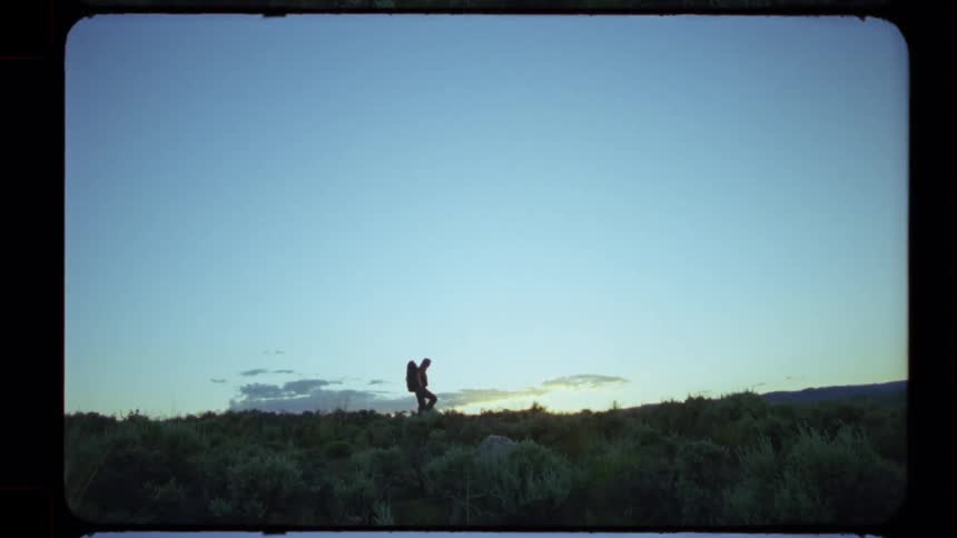 Vintage Super 8 film captures the silhouette of a lone hiker with a backpack walking across a hilltop at sunset