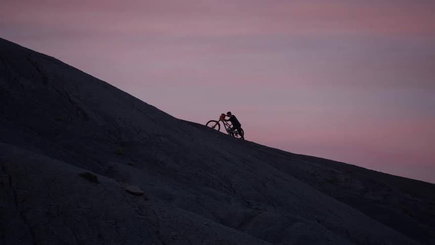 Silhouette of a determined mountain biker struggling to push their bike up a steep mountain at dusk