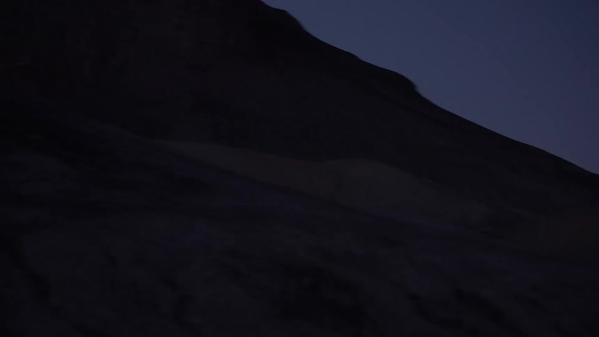 An extreme mountain biker performs tricks on a dark desert trail at dusk, their path illuminated only by a headlamp in this dramatic slow-motion shot