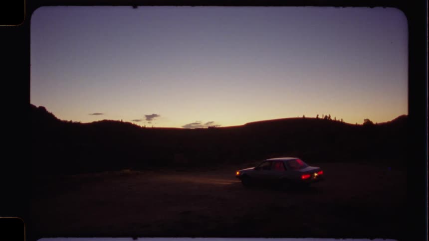 A vintage car with its lights on is parked in a rural landscape at twilight in an authentic film shot