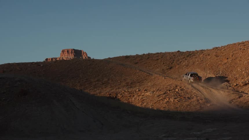 An off-road truck and UTV ascend a winding dirt trail in the vast desert landscape during a beautiful golden hour sunset