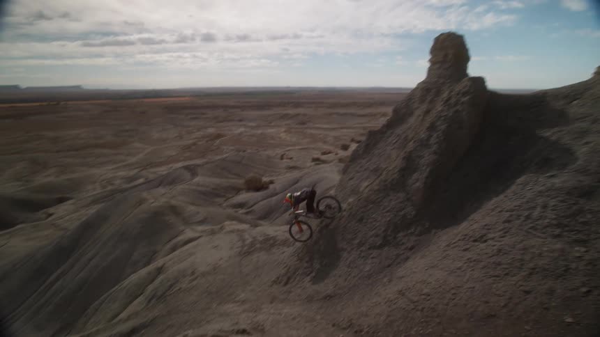 In slow motion, a mountain biker carefully descends a steep and treacherous dirt ridge in a vast badlands landscape