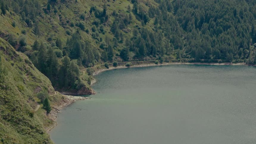 A tranquil mountain lake curves around a lush, tree-covered hillside on a peaceful day