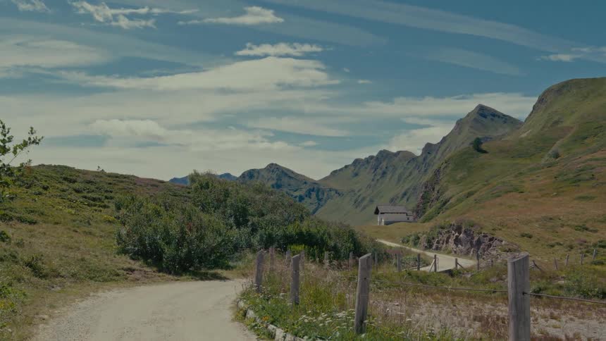 A serene view of a dirt road winding through a lush green mountain valley towards a distant hut