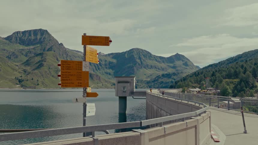 A yellow hiking trail signpost stands on a dam, with a tranquil mountain lake and towering peaks creating a scenic backdrop