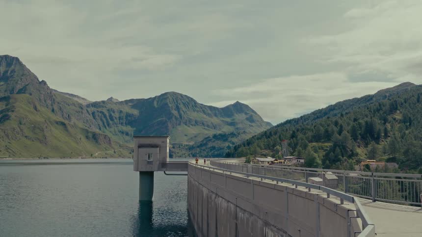 A scenic wide shot of a concrete dam and reservoir nestled in a beautiful green mountain landscape under a cloudy sky