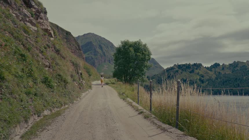 A woman in a yellow skirt walks down a secluded dirt road through the mountains next to a large lake