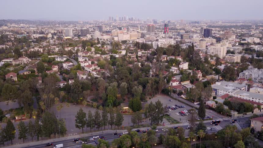 A slow aerial push-in over a lush green park and residential neighborhood in Los Angeles, with the downtown skyline in the background at dusk