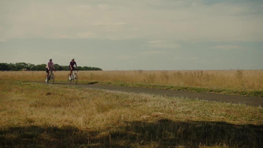 Two women enjoy a scenic bike ride on a rural path through golden fields under a partly cloudy sky
