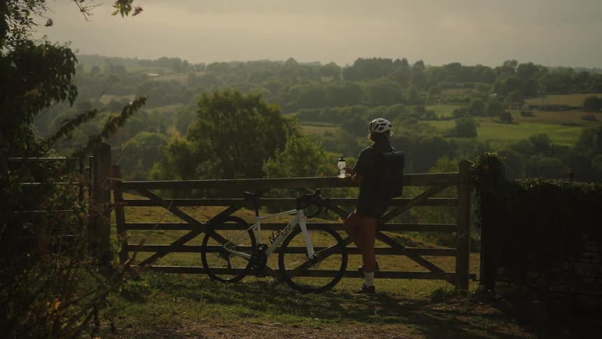 A female cyclist stops at a wooden gate to drink water and enjoy the scenic view of the lush, green countryside