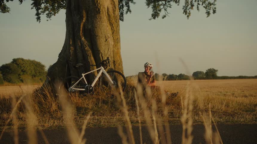A cyclist rests in the shade of a large tree, drinking water in a golden field at sunset
