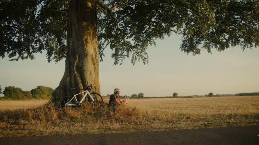 A male cyclist takes a well-deserved rest in the shade of a large tree, enjoying the peaceful countryside view at golden hour
