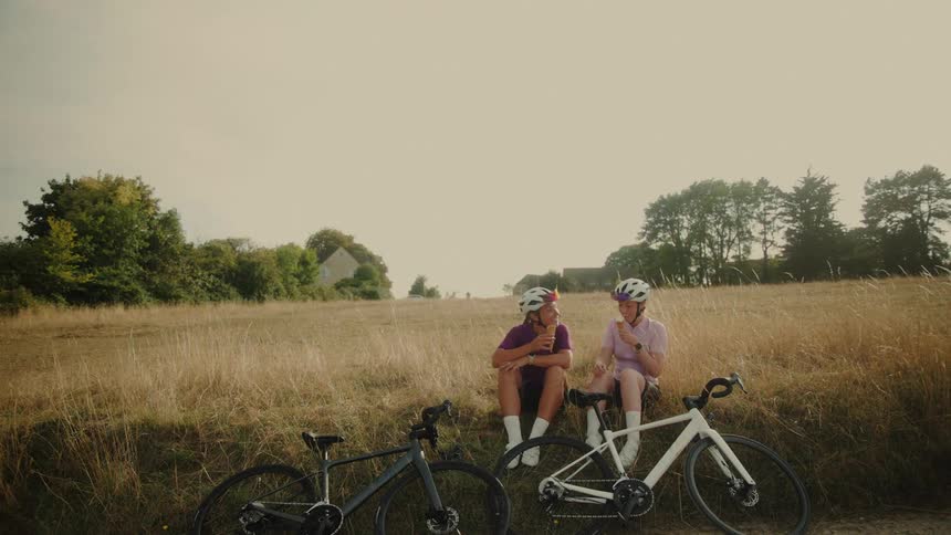 Two female cyclists take a break from their ride, sitting in a sunny field eating ice cream and laughing together