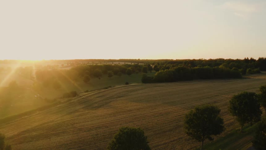 A slow aerial ascent reveals a vast rural landscape with rolling fields and a tree-lined road bathed in the warm light of a golden sunset