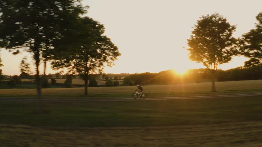 Side-view tracking shot of a cyclist riding a bicycle on a country road with trees during a beautiful golden sunset