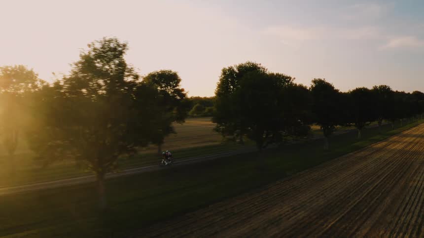 An aerial view follows two cyclists riding along a scenic country road flanked by fields and trees at golden hour