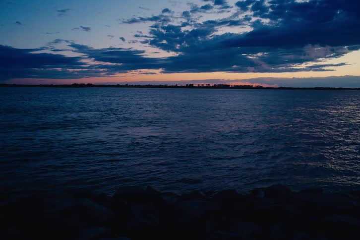 A serene and moody view of a large lake at twilight, with gentle waves and a colorful sunset on the horizon