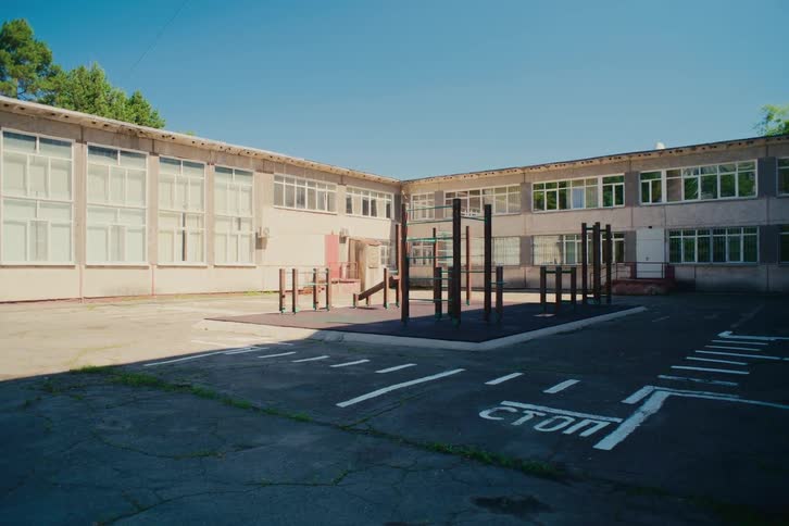 A quiet and empty schoolyard with a small playground and road markings sits peacefully in front of an old school building on a bright sunny day
