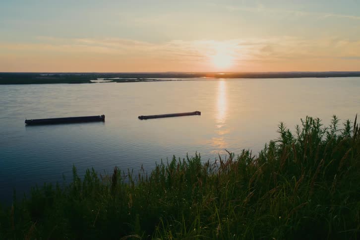 A tranquil sunset over a wide river with two barges peacefully floating as seen from a grassy overlook