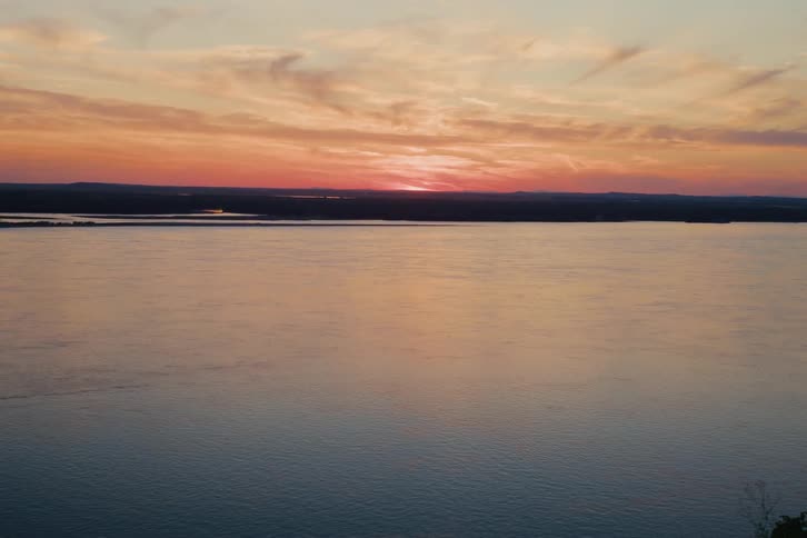 The setting sun casts a warm orange and pink glow over a vast, tranquil river