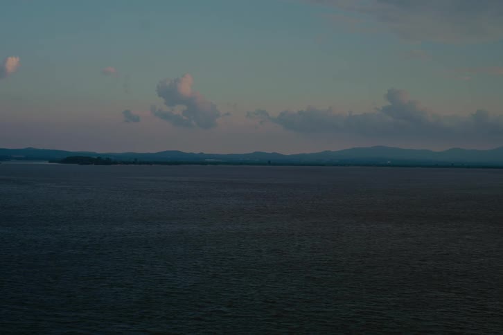 A tranquil and still shot of a vast lake at twilight, with gentle ripples and a soft, colorful sky above the distant shoreline