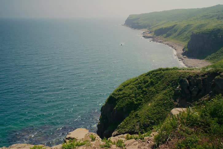 A stunning view from a high cliff overlooking a beautiful, rugged coastline with calm turquoise waters on a hazy day