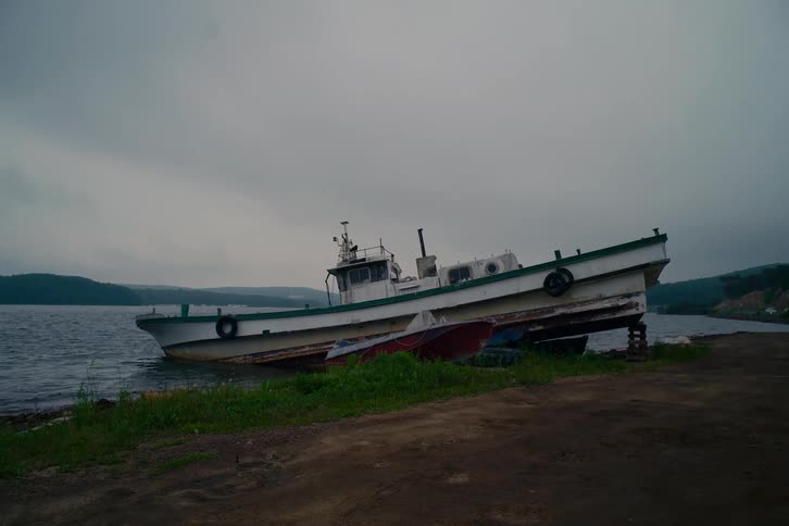 A weathered fishing boat sits beached and abandoned on the shore of a lake on a gloomy, overcast day
