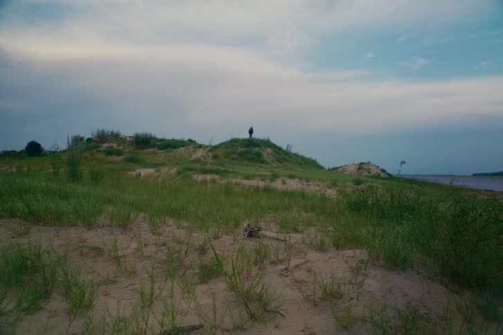 A lone figure walks along the ridge of a grassy sand dune next to the water on an overcast day