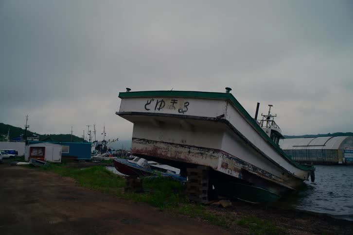 A weathered Japanese fishing boat rests on blocks at the water's edge in a harbor on a gloomy, overcast day