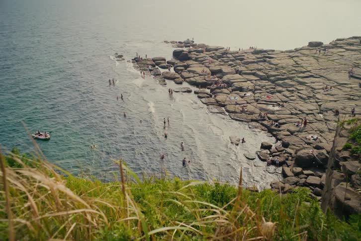 A scenic high-angle view captures people enjoying a summer day swimming and sunbathing along a unique rocky coastline