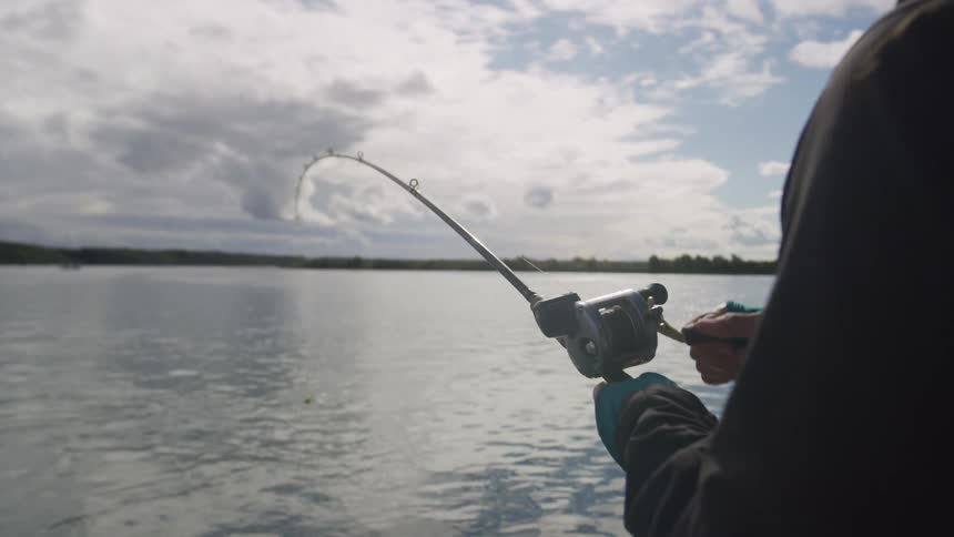 A fisherman reels in a catch on a calm lake under a partly cloudy sky with the sun shining brightly