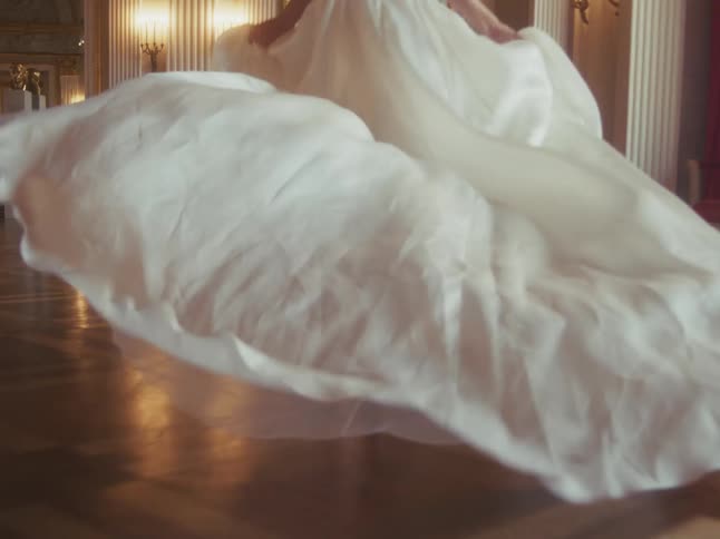 The hem of a flowing white dress swirls in slow motion as a barefoot woman dances across a polished ballroom floor