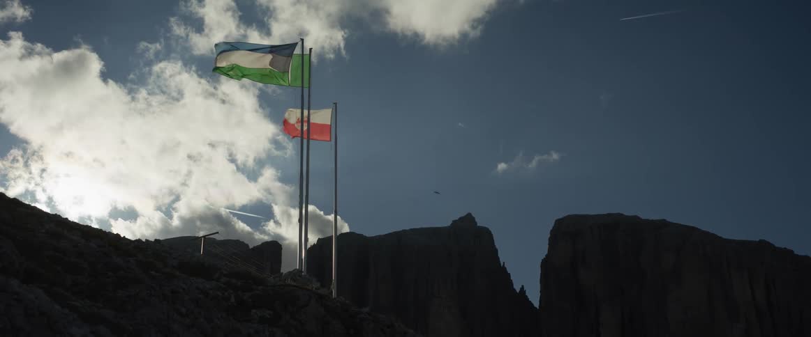 Flags wave in slow motion on a mountaintop, silhouetted against a dramatic sky with a bird flying by