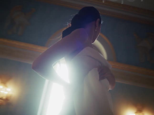 A graceful woman in a flowing white dress dances slowly and elegantly in a grand, sun-drenched hall
