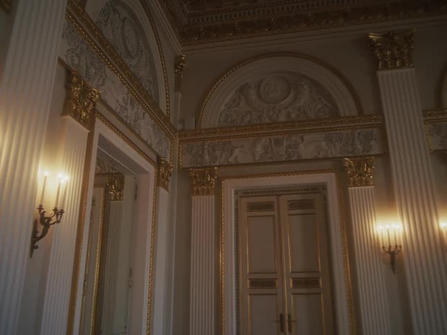 A slow pan across the opulent interior of a classical-style hall featuring white columns, gold leaf details, and ornate wall sconces
