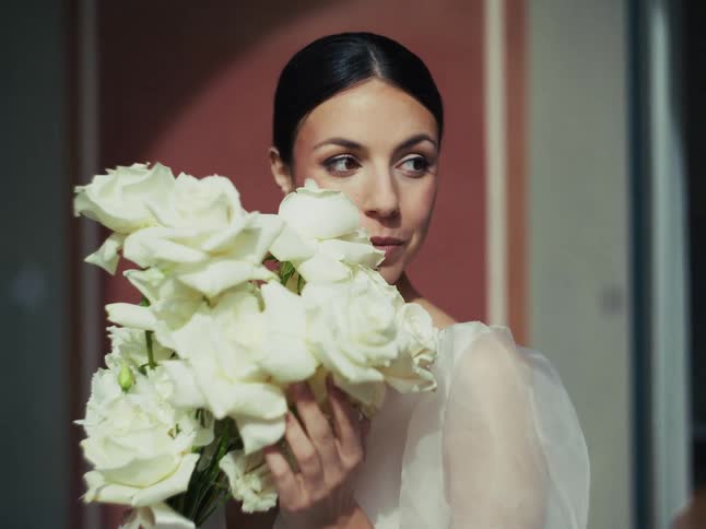 A beautiful bride with dark hair pulled back holds a lovely bouquet of white roses and looks off to the side with a gentle expression