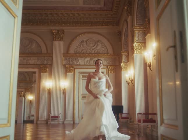 A joyful bride in a flowing white wedding gown runs barefoot towards the camera in slow motion through a magnificent, empty palace ballroom