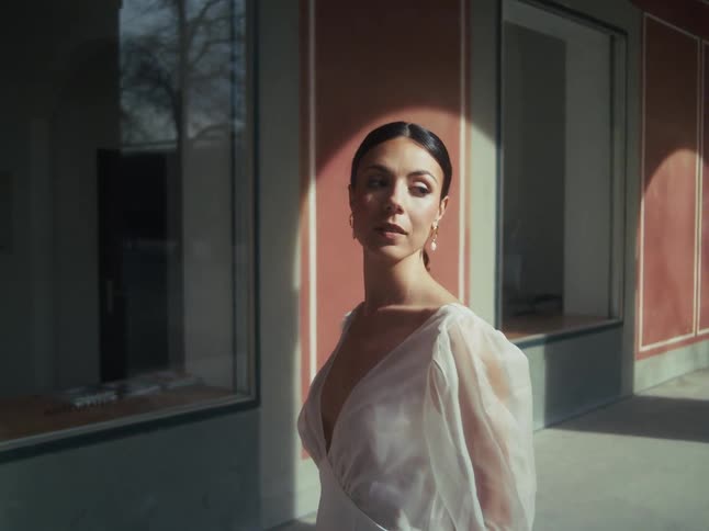 An elegant woman in a white dress stands in a sunlit portico, slowly turning to look into the camera with a sophisticated and direct gaze