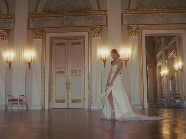 An elegant bride in a beautiful white wedding gown stands alone in a grand, empty ballroom, holding her bouquet
