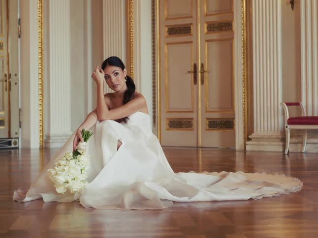 A beautiful bride in an elegant white wedding gown sits pensively on the floor of a luxurious hall with a bouquet of flowers