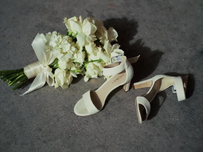 A still life of white wedding heels and a floral bouquet on a concrete floor as a small bee flies around the flowers