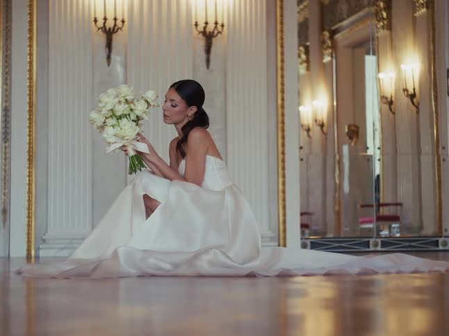 An elegant bride in a stunning white dress with a long train smells her beautiful bouquet in a grand, opulent room