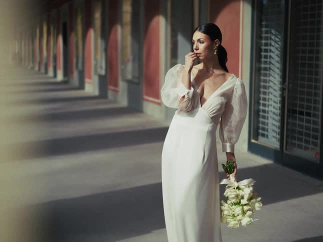 An elegant bride in a stunning white wedding gown holds a bouquet while posing thoughtfully in a long, sun-drenched arched corridor