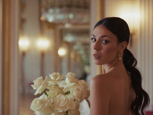 An elegant bride holding a bouquet of white roses turns to look over her shoulder with a serious expression in a luxurious hall