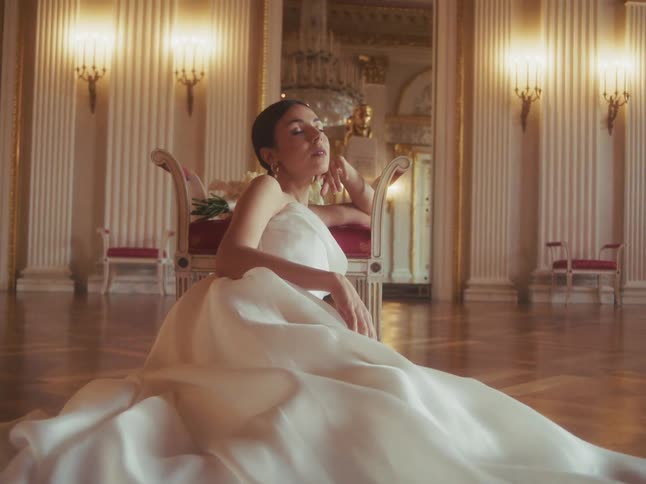 A slow pull-out from the hem of a white wedding dress reveals a beautiful bride sitting serenely on the floor of a grand ballroom