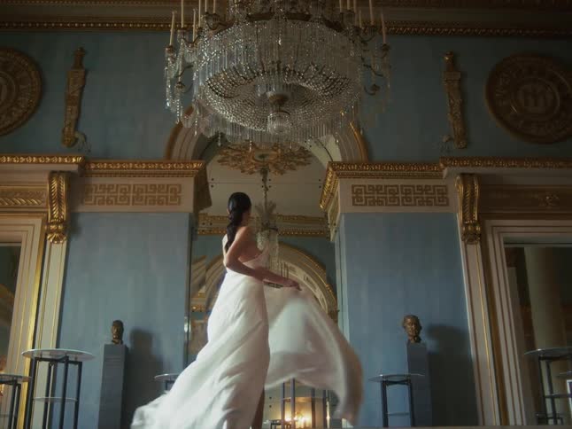 A beautiful woman in a flowing white gown twirls gracefully in slow motion under a crystal chandelier in a luxurious ballroom