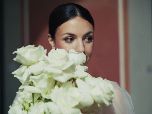 A beautiful bride with her hair in a bun gently smells her large bouquet of white roses on her wedding day