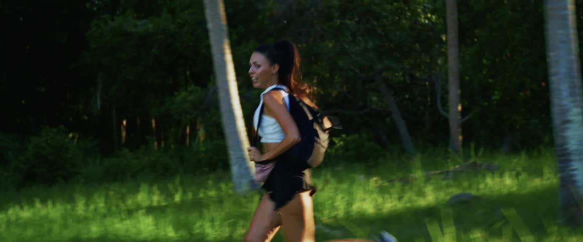 An athletic woman wearing a backpack jogs through a lush green tropical forest