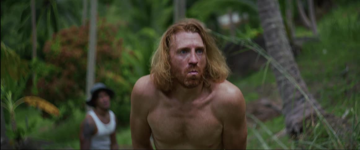 A shirtless man with long red hair eating a chocolate bar in the jungle is suddenly startled and looks around nervously