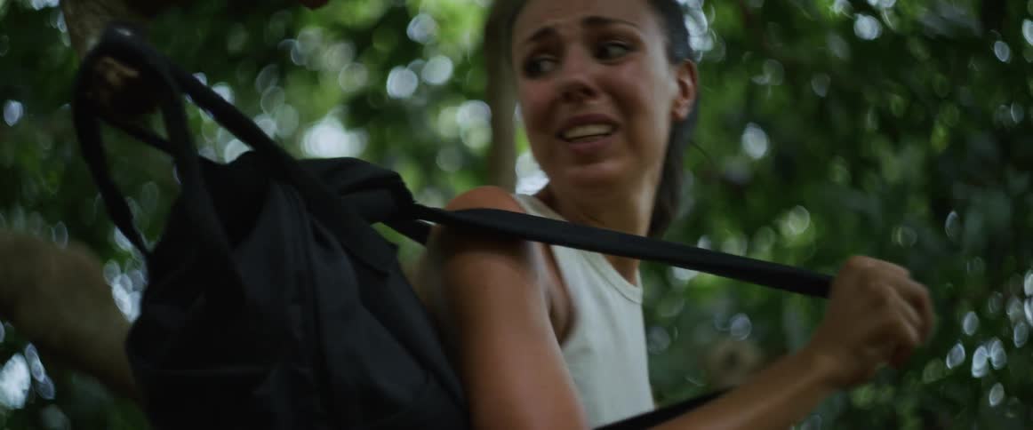 A terrified woman frantically tries to free her backpack snagged on a tree branch while running through a dense jungle